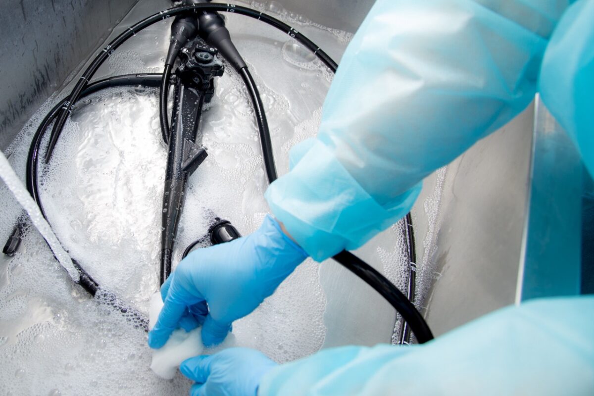 A technician in full PPE washes an endoscope in a sink.